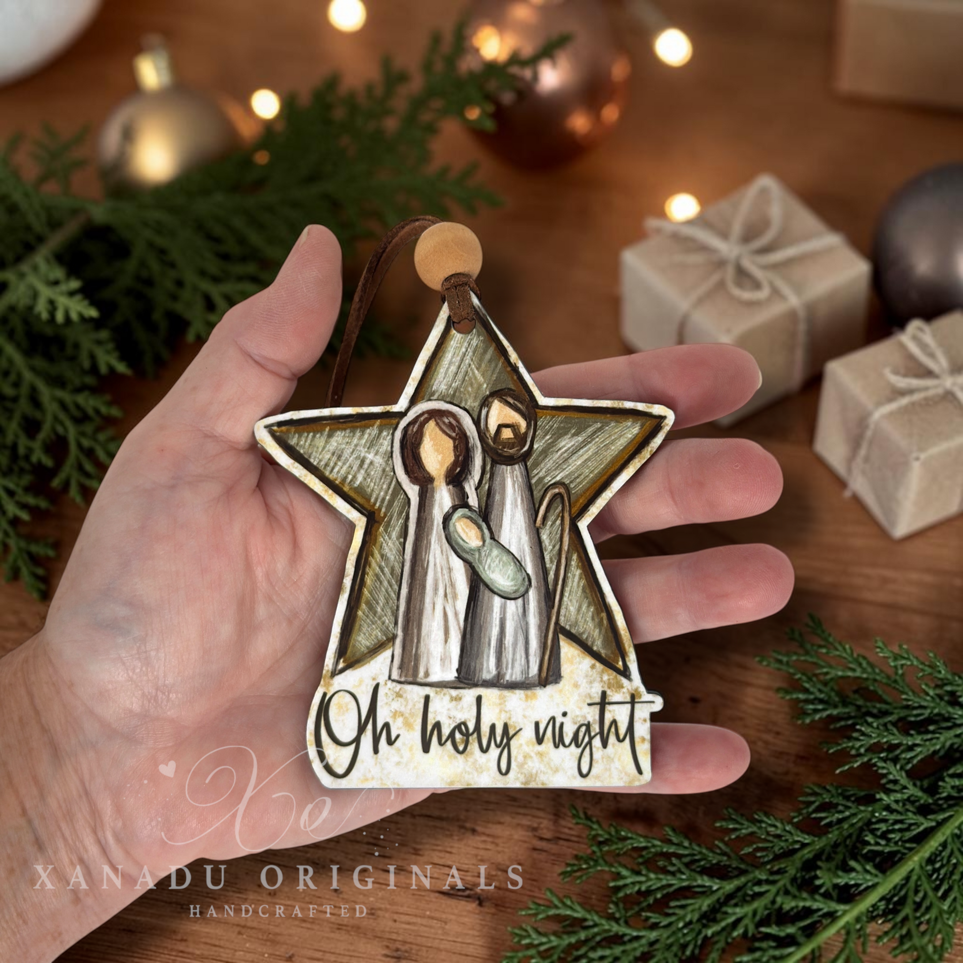 A hand holds a star-shaped Christmas ornament featuring a nativity scene with the text 'Oh holy night' on a rustic wooden surface with holiday decorations in the background.