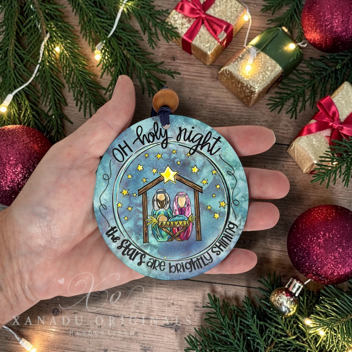 A hand holds a round wooden Christmas ornament featuring a nativity scene with the text 'Oh holy night the stars are brightly shining.' The background is decorated with pine branches, warm string lights, small wrapped gift boxes, and red and gold ornaments.
