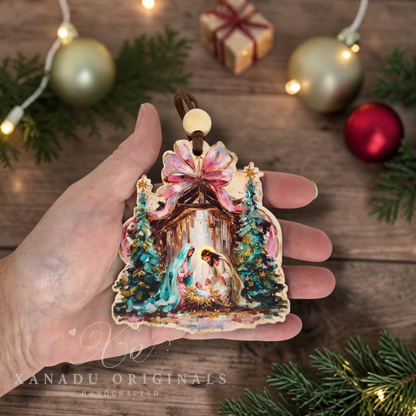 A hand holds a handcrafted nativity ornament featuring Mary, Joseph, baby Jesus in a manger, and decorated trees, with a pink bow at the top. The background shows a wooden surface with Christmas ornaments, pine branches, and warm fairy lights.
