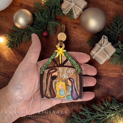 A hand holds a handcrafted Christmas nativity ornament shaped like a stable, decorated with green pine branches, a gold star, and small painted figures. The background shows a wooden surface with gold ornaments, wrapped gifts, and evergreen branches.