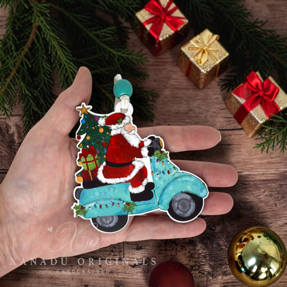 A hand holds a Christmas ornament shaped like Santa Claus riding a light teal scooter, with a decorated tree and gift boxes in the background on a wooden surface. The ornament has a white border and a hanging loop at the top.