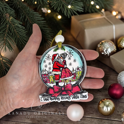 A hand holds a snowglobe shaped Christmas ornament featuring an illustrated Santa Claus and Mrs. Claus inside a snow globe design, with the text 'I saw mommy kissing Santa Claus' on the base. The background shows a festive holiday setting with pine branches, wrapped gifts, and ornaments.