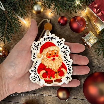 A hand holds a die-cut Christmas ornament featuring an illustrated Santa Claus surrounded by the words 'Believe in the magic of Christmas.' The ornament is shown on a rustic wooden surface with pine branches, ornaments, and wrapped gifts in the background.