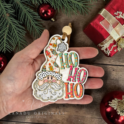 A hand holds a decorative Christmas ornament shaped like Santa's head with the words “HO HO HO” in colorful lettering. The scene is styled with red ornaments, evergreen branches, and a red gift box on a wooden surface.