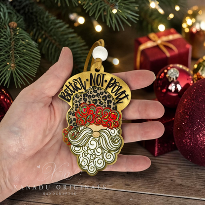 A close-up view of a hand holding the 4-inch leopard print Santa wood ornament to demonstrate its size next to shiny red Christmas ornaments and a wrapped gift box.