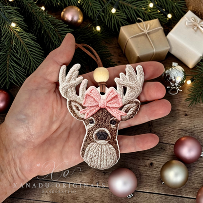 A person holding a 3.75-inch faux knit deer wood ornament in their palm to demonstrate its size, set against a background of wrapped gifts and pink bulbs.