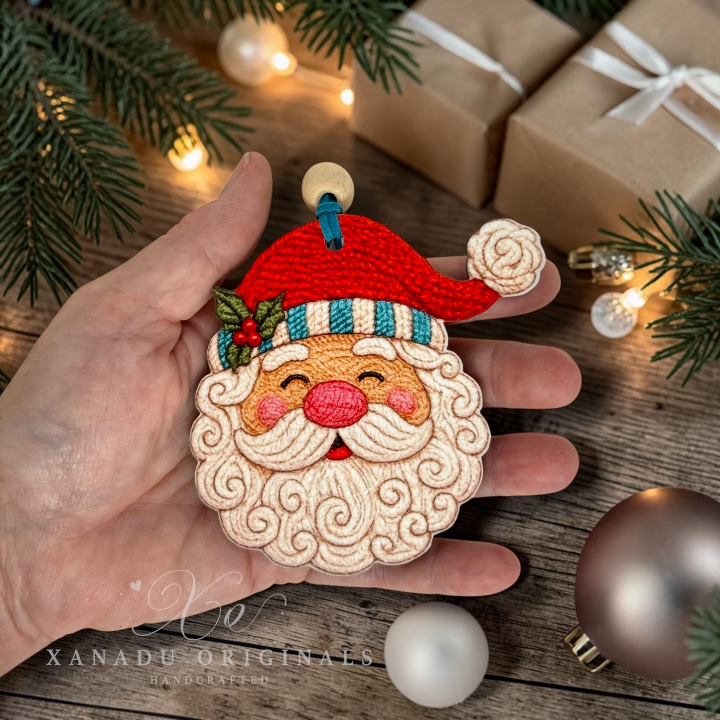 A hand holds a wooden decorative embroidered Christmas ornament shaped like Santa's face. The ornament has a red hat, white beard, pink nose, and green holly accents, photographed on a rustic wooden surface with pine branches, wrapped gifts, and small warm lights in the background.