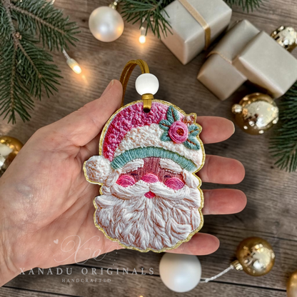A hand holds a Santa Claus embroidered ornament with a gold-tone hanger and white bead. The ornament features pink, white, and teal embroidery with a small pink flower.
