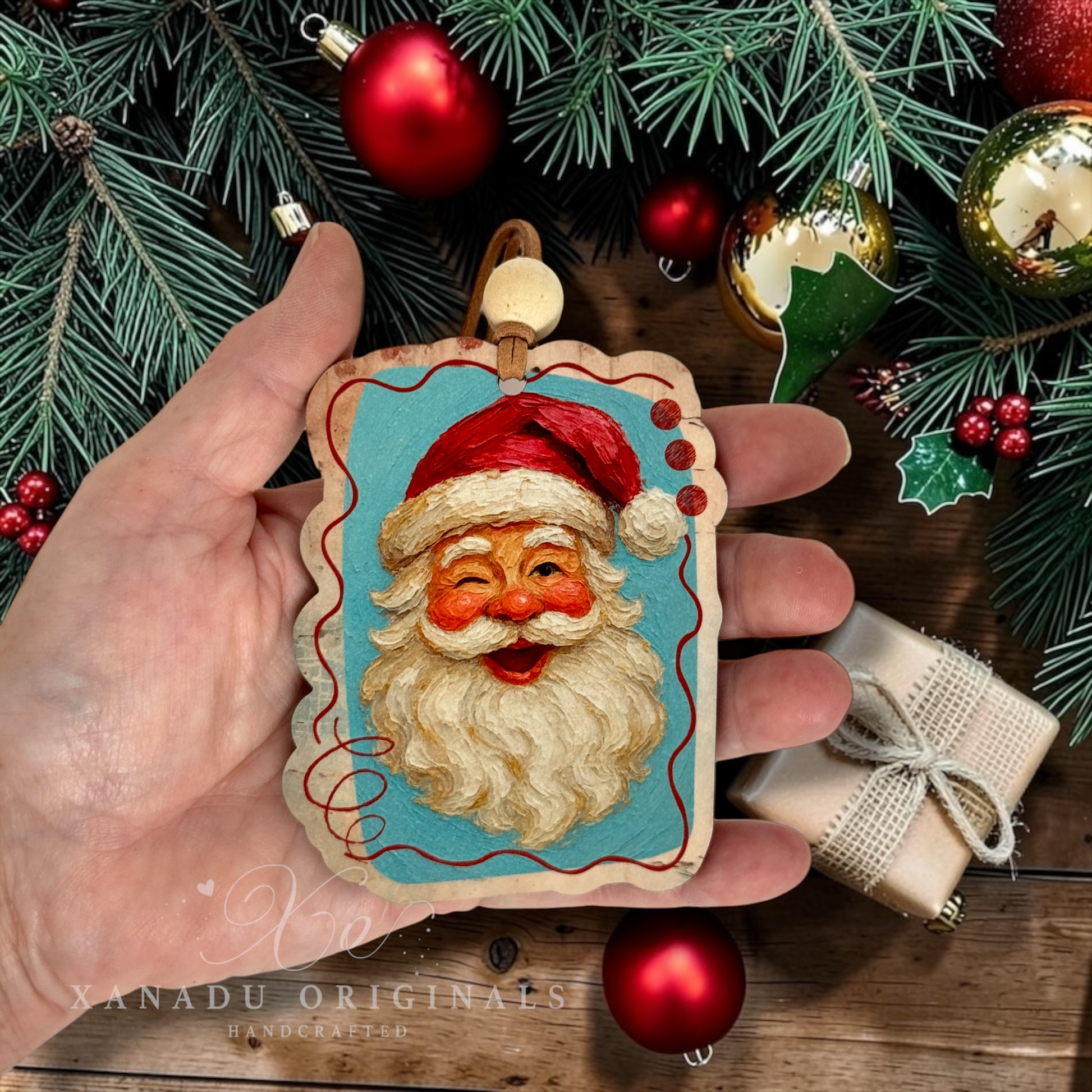 A hand holds a rectangular Santa Claus ornament with a turquoise background, red hat, and white beard, surrounded by Christmas decorations including red ornaments, pine branches, berries, and a wrapped gift. A watermark reading XANADU ORIGINALS is visible at the bottom.