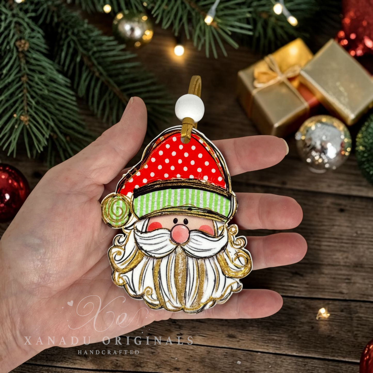 A hand holds a handcrafted Christmas ornament shaped like Santa's face, featuring a red hat with white polka dots, a green striped band, a white beard with gold glitter accents, and a pink nose. The background shows a rustic wooden surface with pine branches, holiday lights, and wrapped gifts.