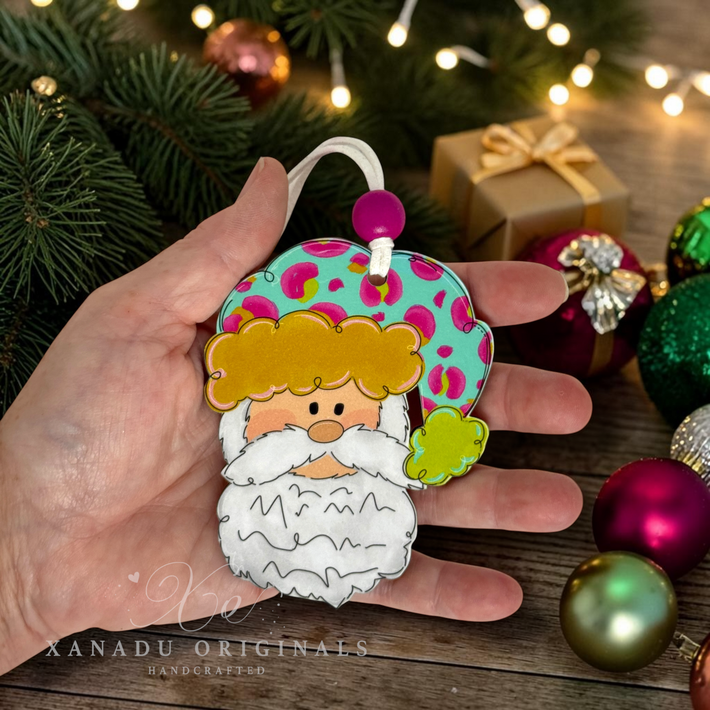 A hand holds a colorful Christmas ornament shaped like a stylized Santa face with a white beard, yellow hair, and a teal hat with pink spots. The background shows holiday greenery, ornaments, and warm string lights, with a watermark reading XANADU ORIGINALS.