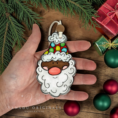 A person holding a dark skin tone Santa wood ornament in their palm, showing its size alongside red and green metallic Christmas bulb decorations and a small red gift box.