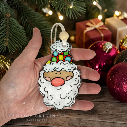 A hand holding a light skin tone Santa Claus wood ornament to demonstrate its 4.25-inch height, surrounded by festive holiday decorations, shiny pink bulbs, and a red gift package.