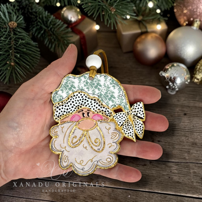 A hand holds a handcrafted Christmas ornament shaped like a Santa face with a green patterned hat, white beard, black-and-white polka dot pattern, and gold trim, set against a rustic holiday background with pine branches, ornaments, and wrapped gifts.