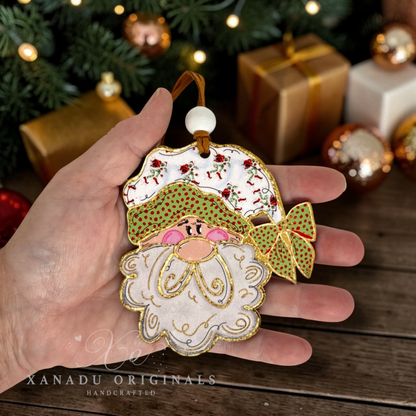 A hand holds a handcrafted Christmas ornament shaped like a Santa-style face with a white beard, green hat with red dots, pink nose, gold trim, and a green bow. The background shows a festive holiday scene with wrapped gifts, ornaments, and Christmas tree branches.