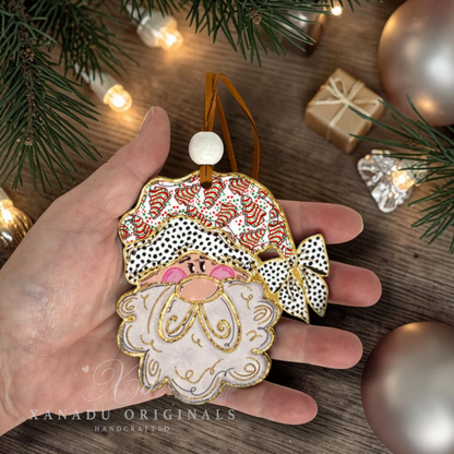 A hand holds a handcrafted Christmas ornament shaped like Santa's face, decorated with patterned fabric, gold trim, and a white beard, set against a rustic holiday background with pine branches, lights, and ornaments.