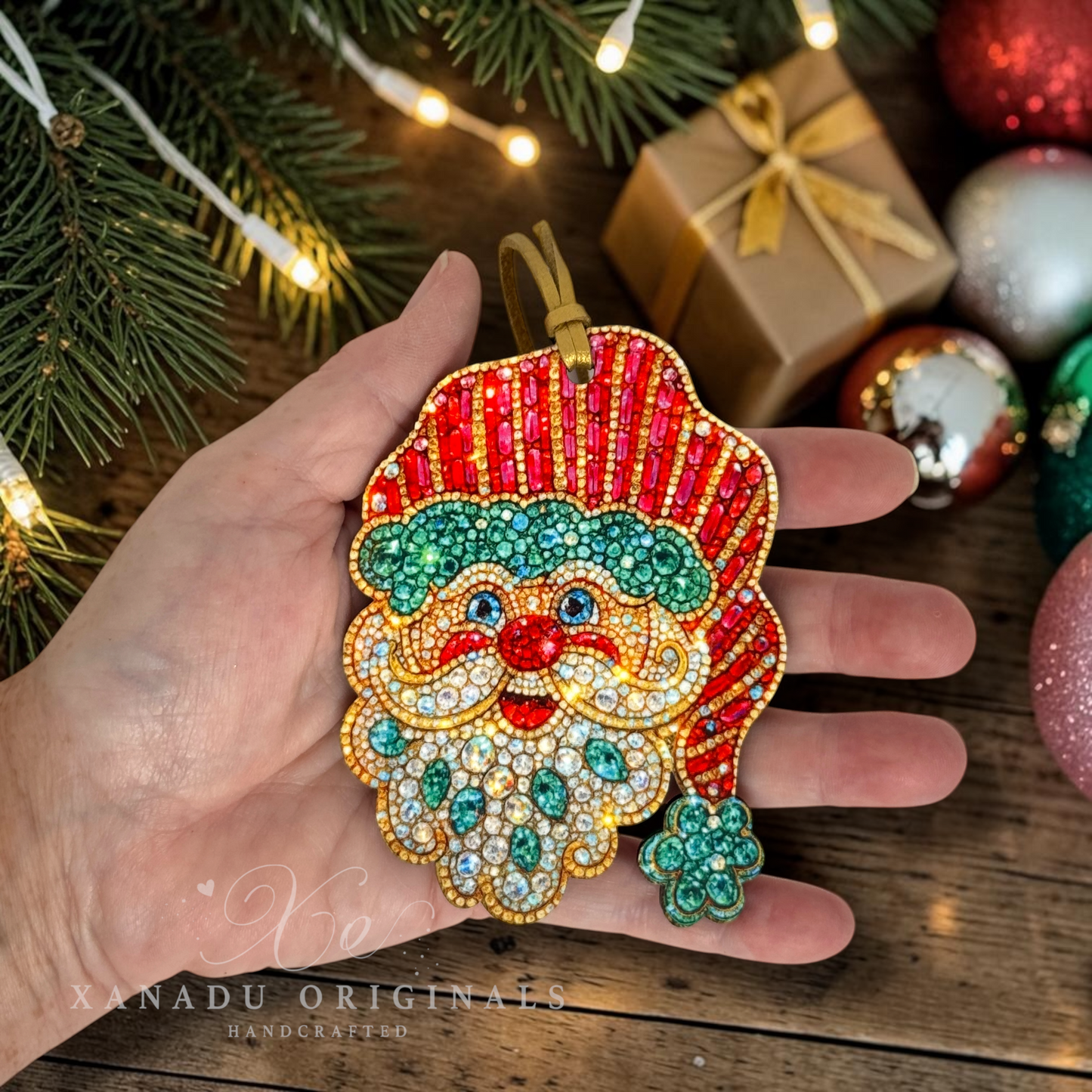 A hand holds a festive Santa Claus ornament decorated with red, green, and gold rhinestones, set against a holiday background with pine branches, wrapped gifts, and other ornaments.