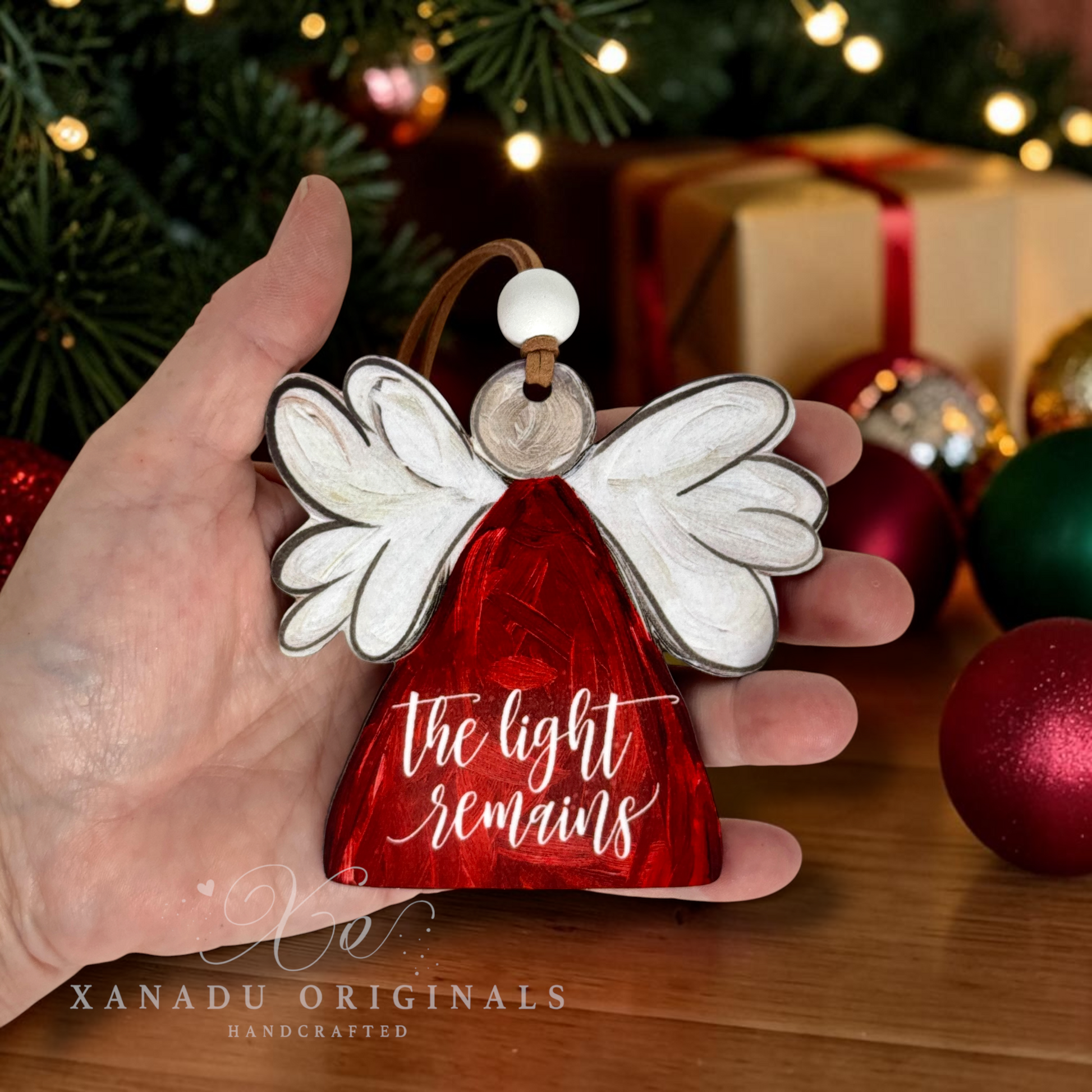 A hand holds a red and white angel Christmas ornament with the words 'the light remains' written on it, set against a festive background of a Christmas tree, wrapped gifts, and ornaments.