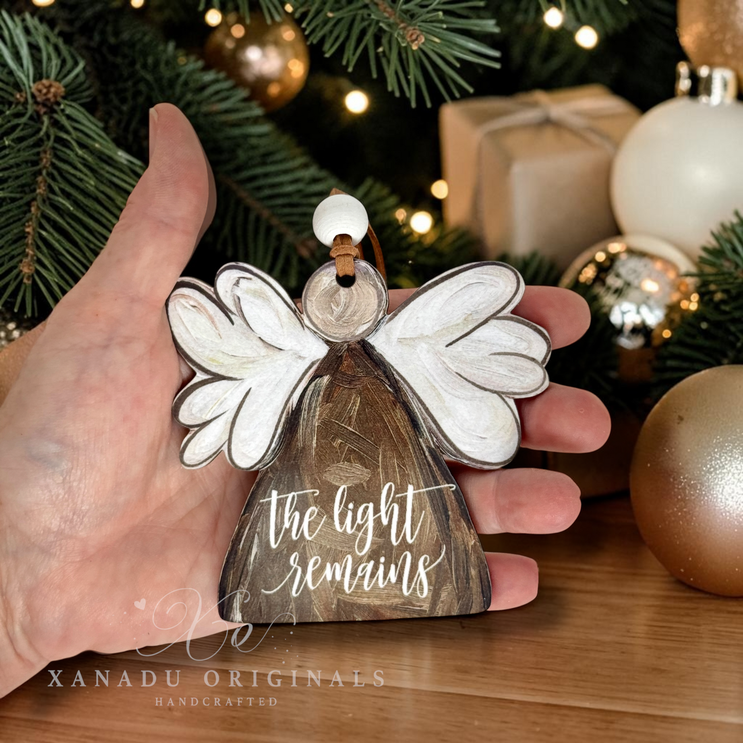 A hand holds a wooden angel Christmas ornament with white wings and the phrase 'the light remains' in white script on a brown body. The background shows a decorated Christmas tree with lights, ornaments, and wrapped gifts.