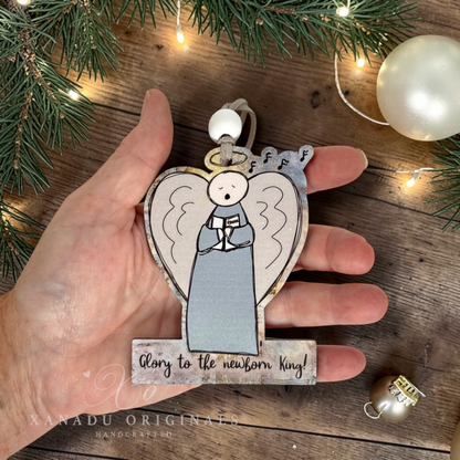 A hand holds a decorative wooden Christmas ornament shaped like an angel with the words 'Glory to the newborn king!' on a rustic wooden surface with evergreen branches and warm string lights in the background.
