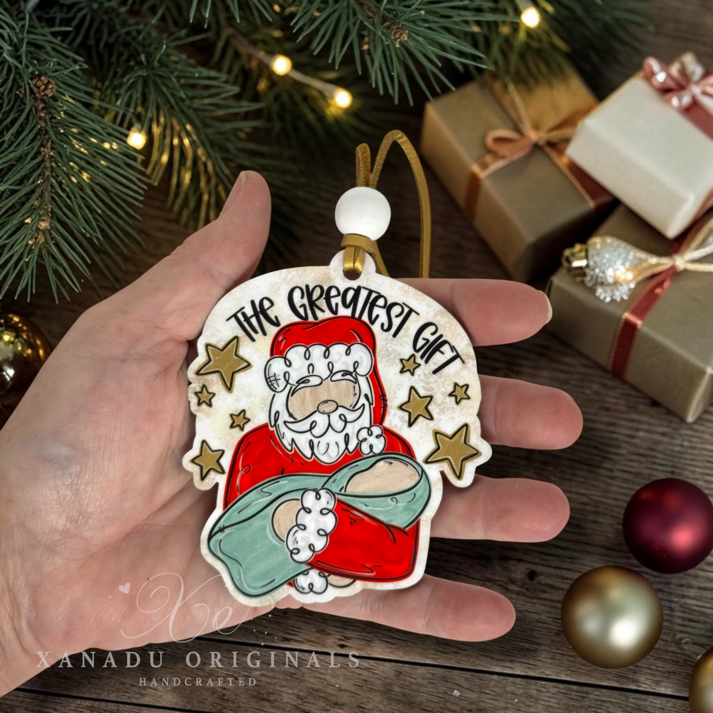 A hand holds a handcrafted Christmas ornament, featuring a painted Santa holding baby Jesus with the text 'THE GREATEST GIFT' and gold stars. The background shows a rustic wooden table with pine branches, wrapped gift boxes, and holiday ornaments.