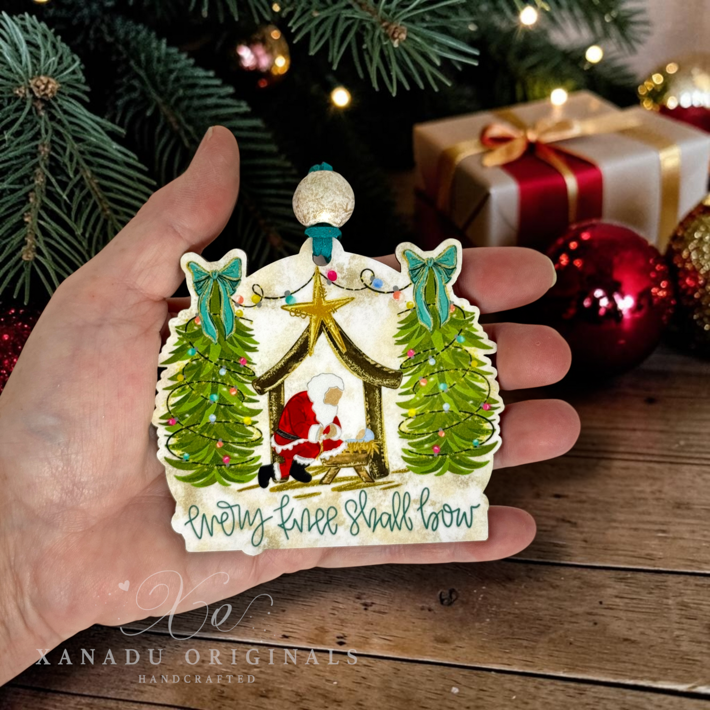 A hand holds a handcrafted Christmas ornament shaped like a nativity scene. The ornament features a cream-colored base with painted green trees, a gold star, a Santa figure, and the text 'Every knee shall bow' at the bottom.