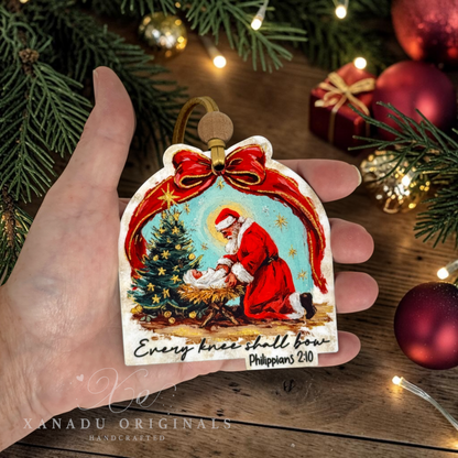 A hand holds a hand-carved Christmas ornament featuring a nativity scene with Santa, baby Jesus, and a tree, with the text 'Every knee shall bow Philippians 2:10'. The background shows a wooden surface with holiday decorations like pine branches, red ornaments, and wrapped gifts.