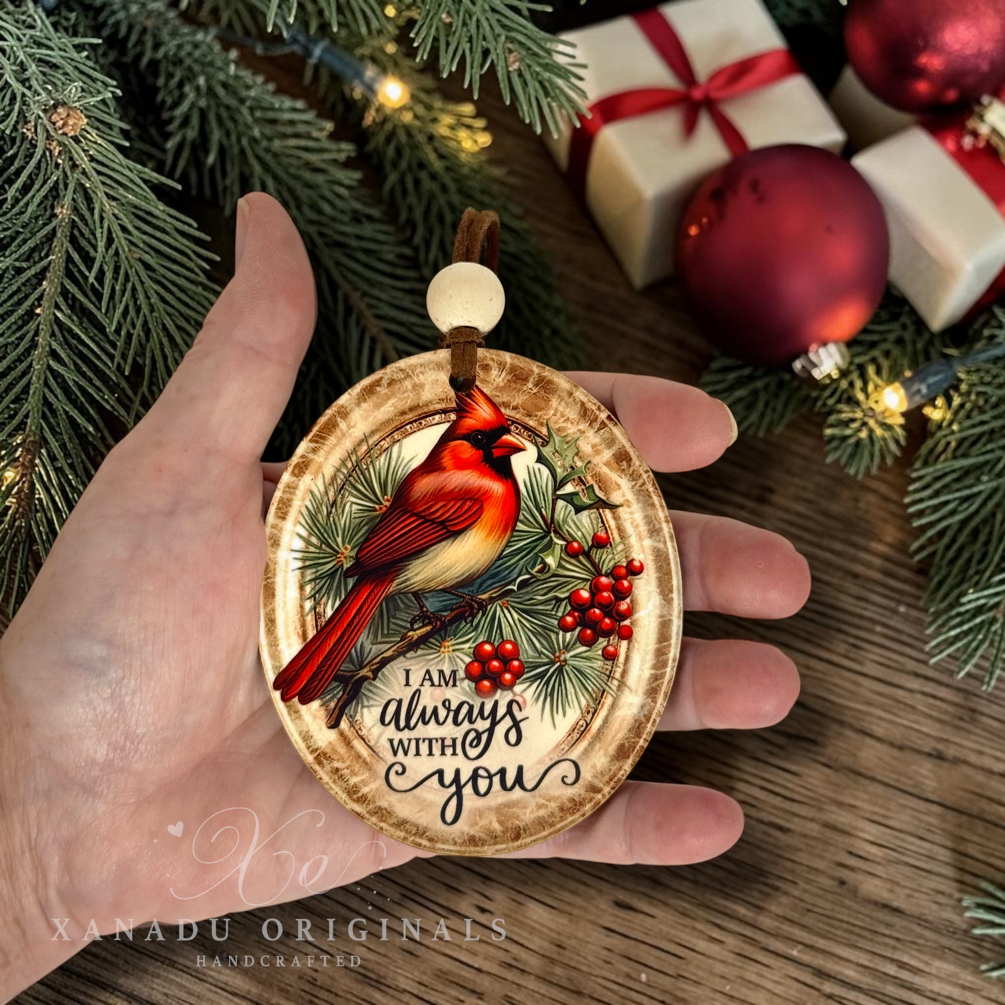 A hand holds an oval Christmas ornament featuring a red cardinal perched on a branch with pine needles and red berries, with the text 'I AM always WITH you.' The background shows evergreen branches, warm string lights, wrapped gift boxes, and red ornaments on a wooden surface.
