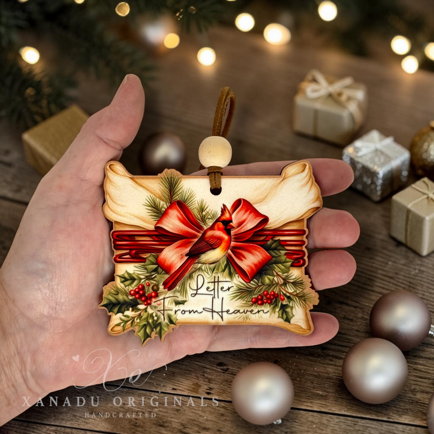 A hand holds a decorative Christmas ornament shaped like a vintage letter, featuring a red cardinal, large red bow, holly, pinecones, and the words 'Leter From Heaven.' The background shows a rustic wooden surface with blurred gift boxes and warm string lights.