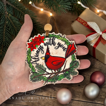 A hand holds a wooden Christmas ornament shaped like a bird perched on a wreath, with the phrase “Always with you” and red poinsettia flowers. The scene includes pine branches, warm string lights, a wrapped gift, and decorative baubles on a rustic wooden surface.