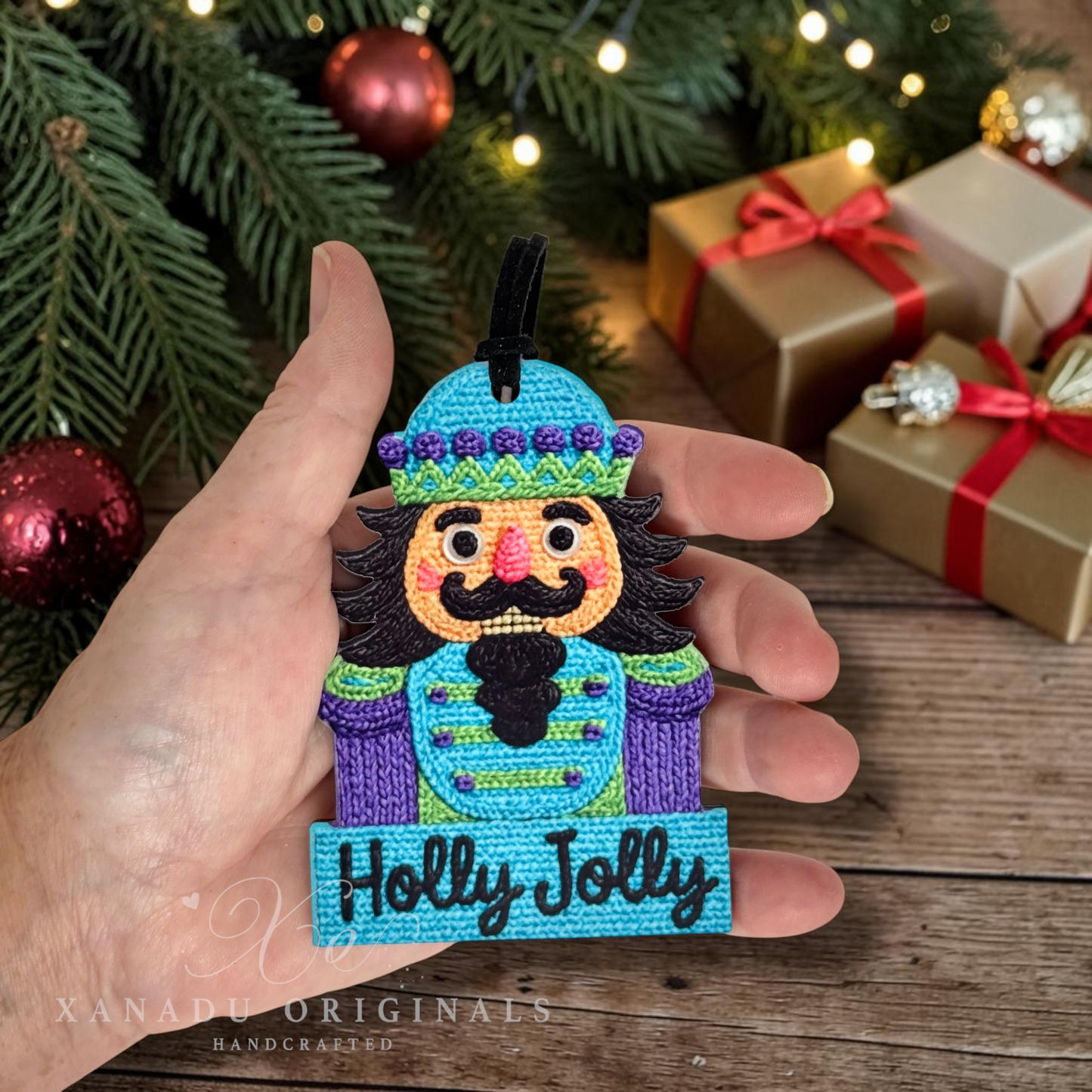 A hand holds a colorful crocheted nutcracker ornament with the words 'Holly Jolly' on a blue banner, set against a festive holiday background with pine branches, ornaments, and wrapped gifts.