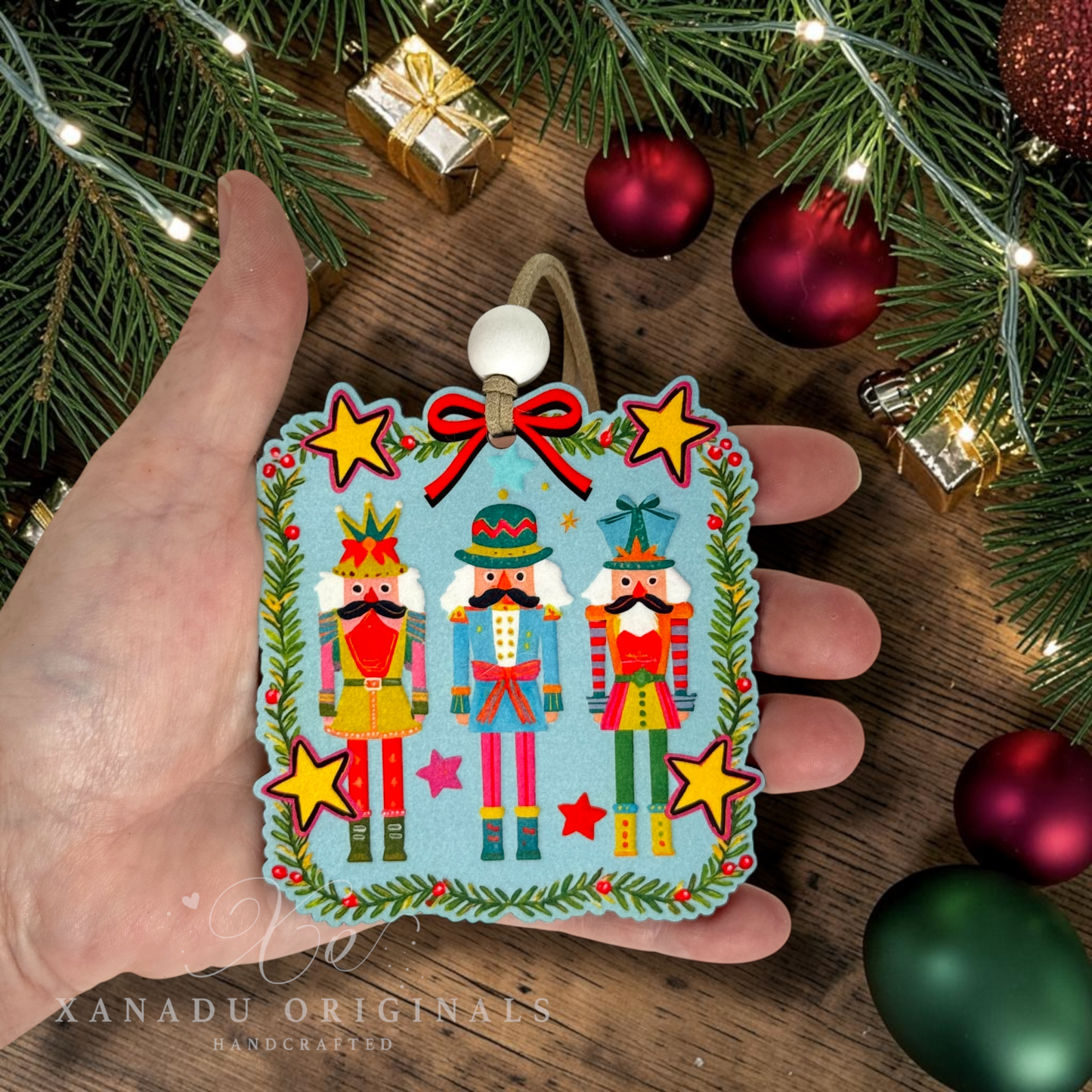 A hand holds a square felt Christmas ornament featuring two nutcracker figures with a red bow and gold stars, surrounded by pine branches, ornaments, and warm string lights. The brand name XANADU ORIGINALS is visible in the bottom left corner.