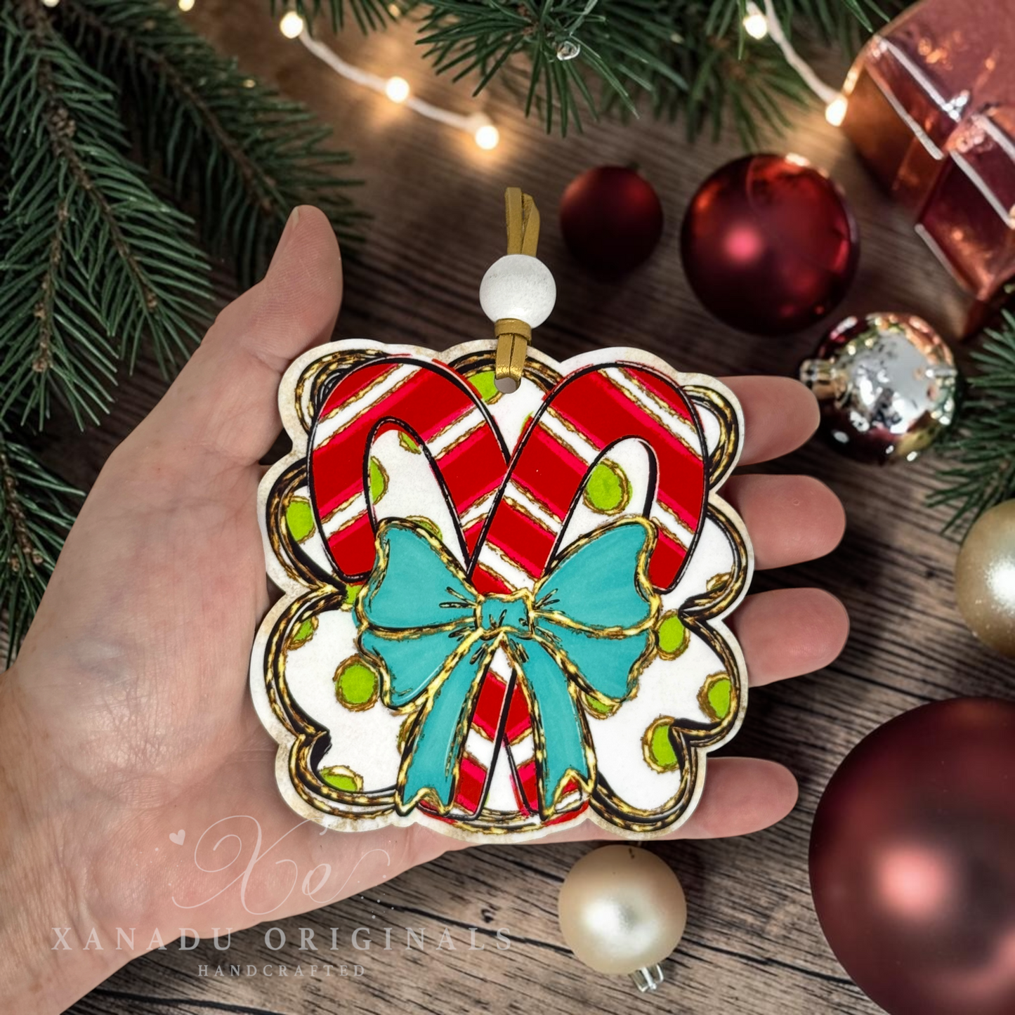 A hand holds a hand-painted wooden Christmas ornament shaped like two candy canes with a turquoise bow. The ornament is decorated with red and white stripes, gold glitter outlines, and small green dots, set against a festive holiday background with pine branches, lights, and other ornaments.