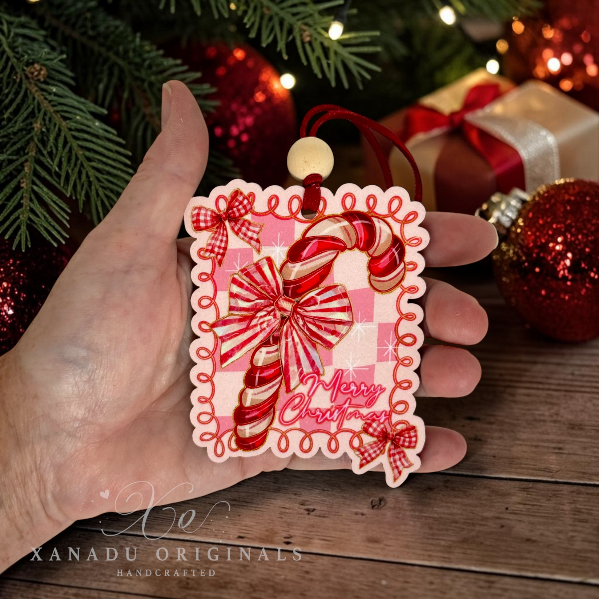 A hand holds a handcrafted Christmas ornament featuring a red and white striped candy cane on a pink gingham background with red ribbon bows and the words 'Merry Christmas.' The scene includes festive green pine branches, red ornaments, and wrapped gifts in the background.