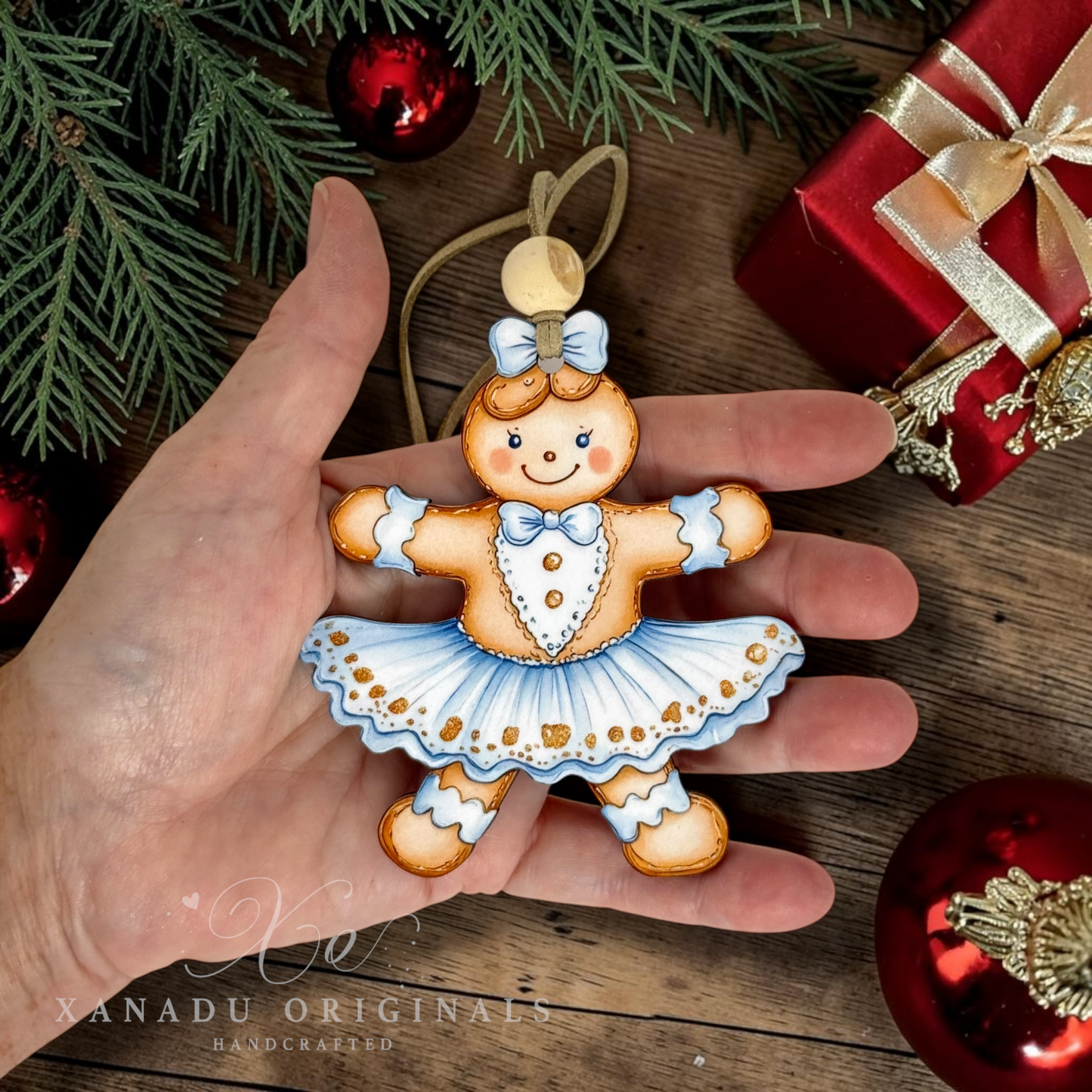 A hand holds a hand-painted gingerbread girl ornament wearing a blue and white tutu dress. The background features a rustic wooden surface decorated with pine branches, red ornaments, and a red gift box.