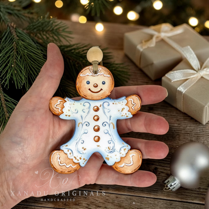 A hand holds a handcrafted gingerbread man Christmas ornament with a light blue icing pattern and gold accents, set against a rustic holiday background with pine branches, wrapped gifts, and a silver ornament.