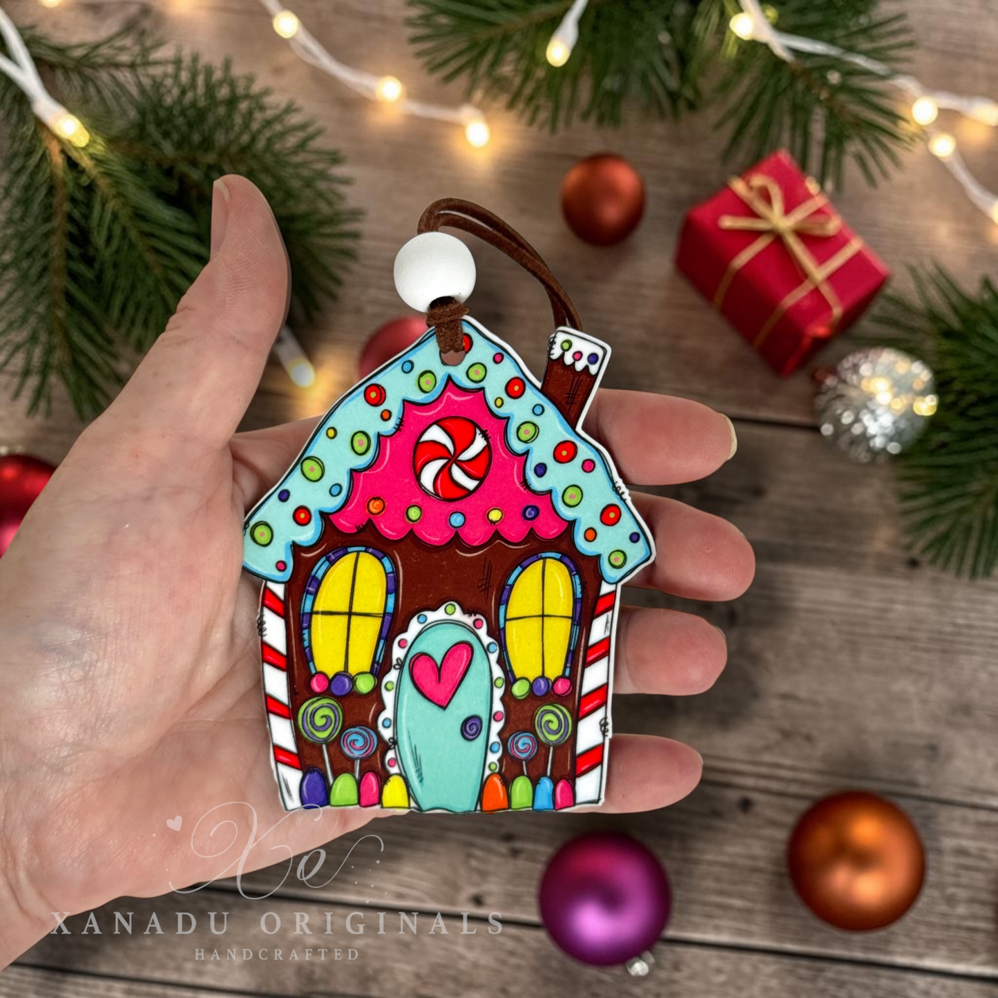 A hand holds a colorful gingerbread house ornament decorated with candy motifs, set against a festive holiday background with pine branches, string lights, ornaments, and a red gift box.