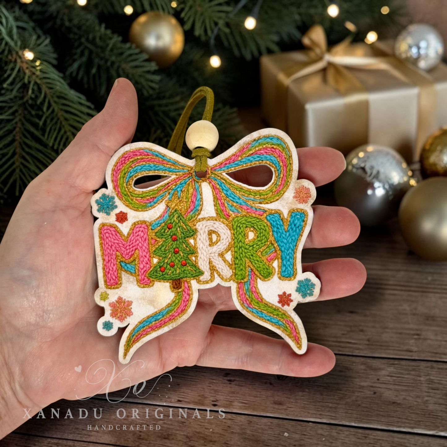 A hand holds a handcrafted embroidered Christmas ornament shaped like a bow and the word “MERRY,” decorated with colorful thread and a small pine tree motif, with a Christmas tree and wrapped gift in the background.