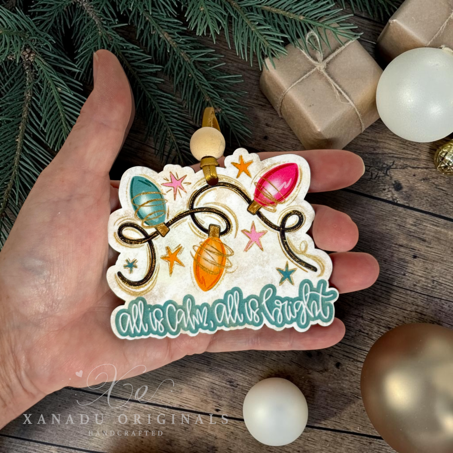 A hand holds a die-cut Christmas ornament featuring colorful holiday string lights and the phrase 'all is calm, all is bright' in teal script. The scene is styled with pine branches, wrapped gift boxes, and ornaments on a rustic wooden surface.