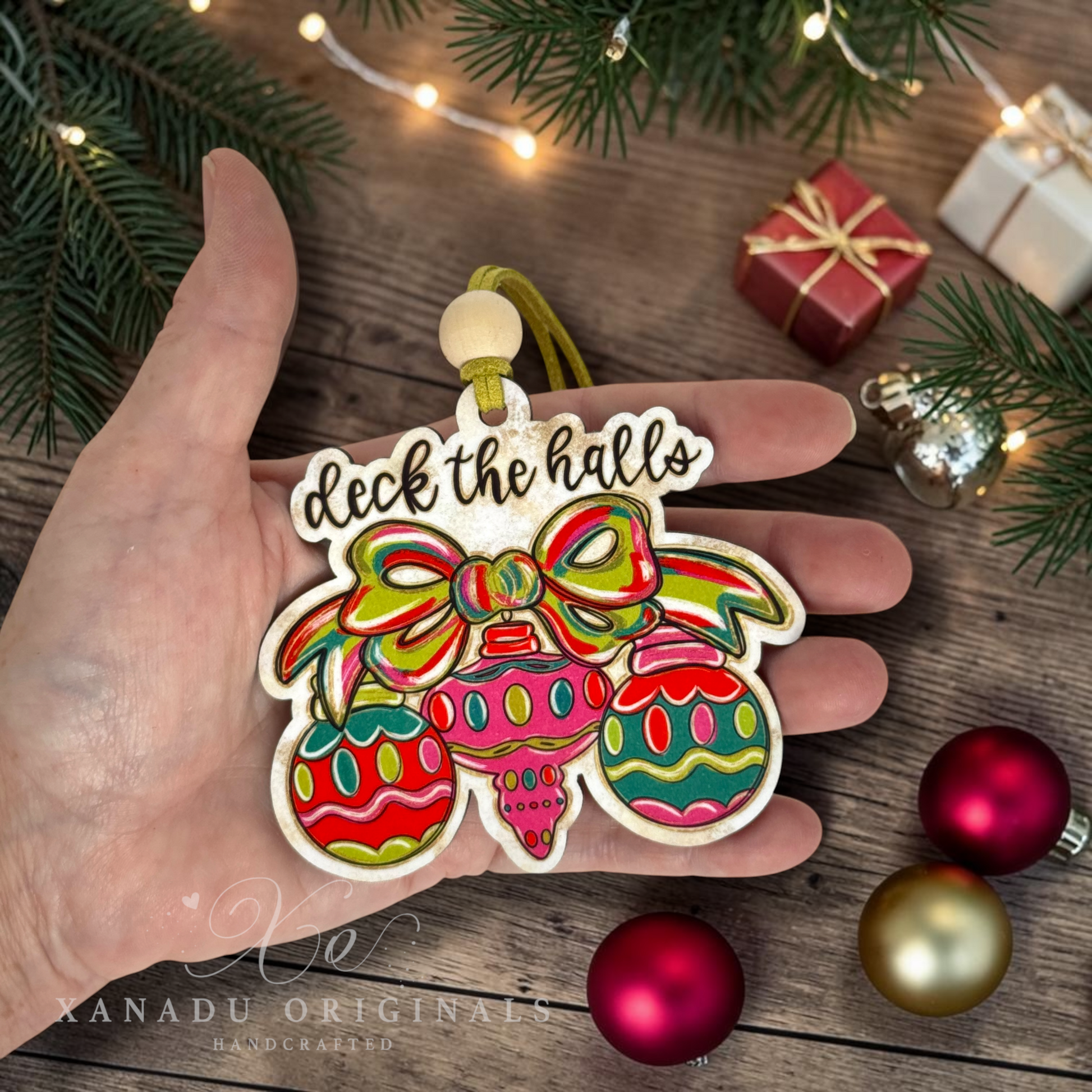 A hand holds a colorful Christmas ornament shaped like a decorated tree with the words “deck the halls” at the top. The scene is styled with pine branches, string lights, small wrapped gifts, and shiny Christmas ornaments on a wooden surface.