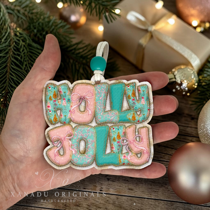 A hand holds a decorative die-cut ornament spelling “HOLLY JOLLY” in pastel pink and turquoise icing with gold glitter accents, surrounded by holiday greenery, ornaments, and wrapped gifts.