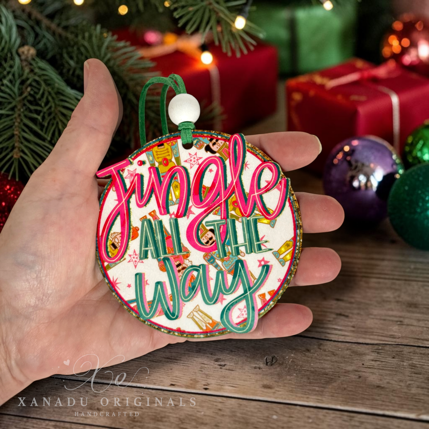 A hand holds a round Christmas ornament that reads 'Jingle All The Way' in pink and green script over a white background with holiday illustrations. The scene is decorated with pine branches, wrapped gifts, and colorful ornaments, with a watermark for XANADU ORIGINALS at the bottom.