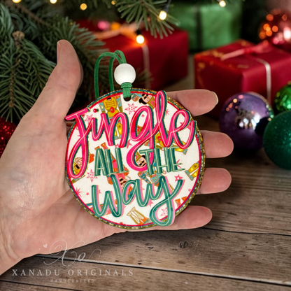 A hand holds a round Christmas ornament that reads 'Jingle All The Way' in pink and green script over a white background with holiday illustrations. The scene is decorated with pine branches, wrapped gifts, and colorful ornaments, with a watermark for XANADU ORIGINALS at the bottom.