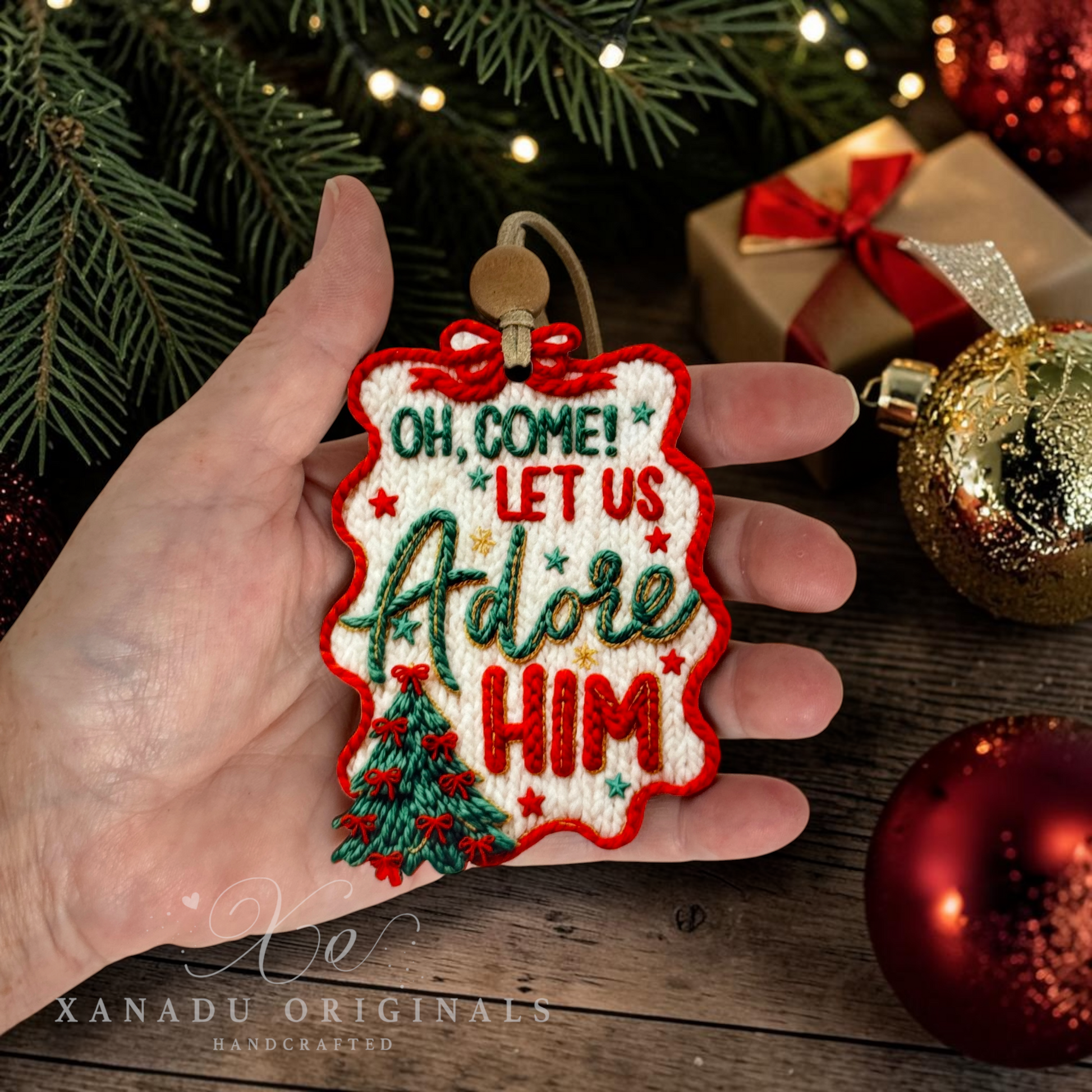 A hand holds a Christmas embroidered ornament that reads 'OH COME! LET US Adore HIM' with a small tree and red border, surrounded by holiday decorations and wrapped gifts on a wooden surface.