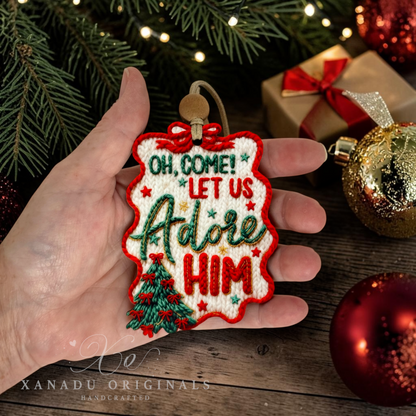 A hand holds a Christmas embroidered ornament that reads 'OH COME! LET US Adore HIM' with a small tree and red border, surrounded by holiday decorations and wrapped gifts on a wooden surface.