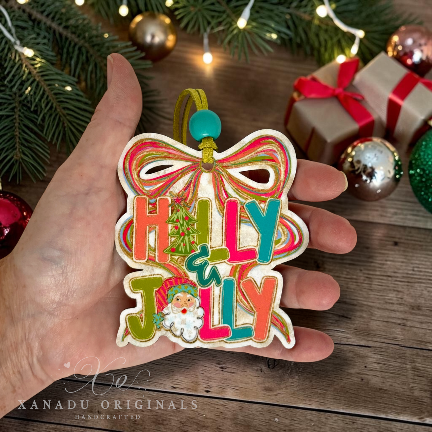 A hand holds a colorful wooden Christmas ornament shaped like a bow with the words “HOLLY JOLLY” in multicolored letters and a small Santa face. The background shows holiday decorations, including pine branches, wrapped gifts, and ornaments, on a wooden surface.
