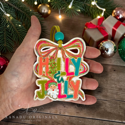 A hand holds a colorful wooden Christmas ornament shaped like a bow with the words “HOLLY JOLLY” in multicolored letters and a small Santa face. The background shows holiday decorations, including pine branches, wrapped gifts, and ornaments, on a wooden surface.