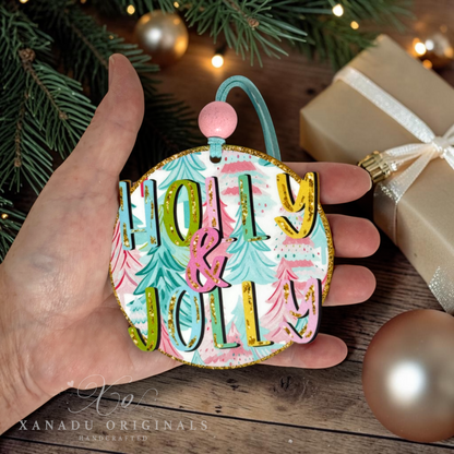 A hand holds a round, handcrafted Christmas ornament featuring the words 'HOLLY & JOLLY' in colorful raised letters over a pastel tree pattern. The ornament has a gold glitter border, a pink hanging bead, and a teal ribbon loop.