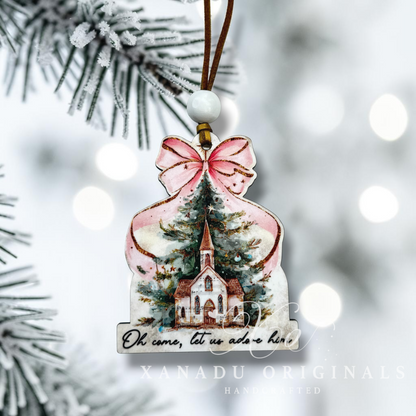 Christmas ornament with a church design and pink bow, hanging on a branch with blurred lights in the background.