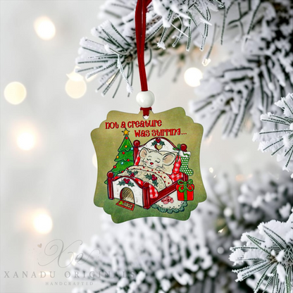 A handcrafted Christmas ornament featuring a cartoon cat sleeping in bed with a tree and gifts, hanging from a red ribbon on a snowy Christmas tree branch with warm bokeh lights in the background.